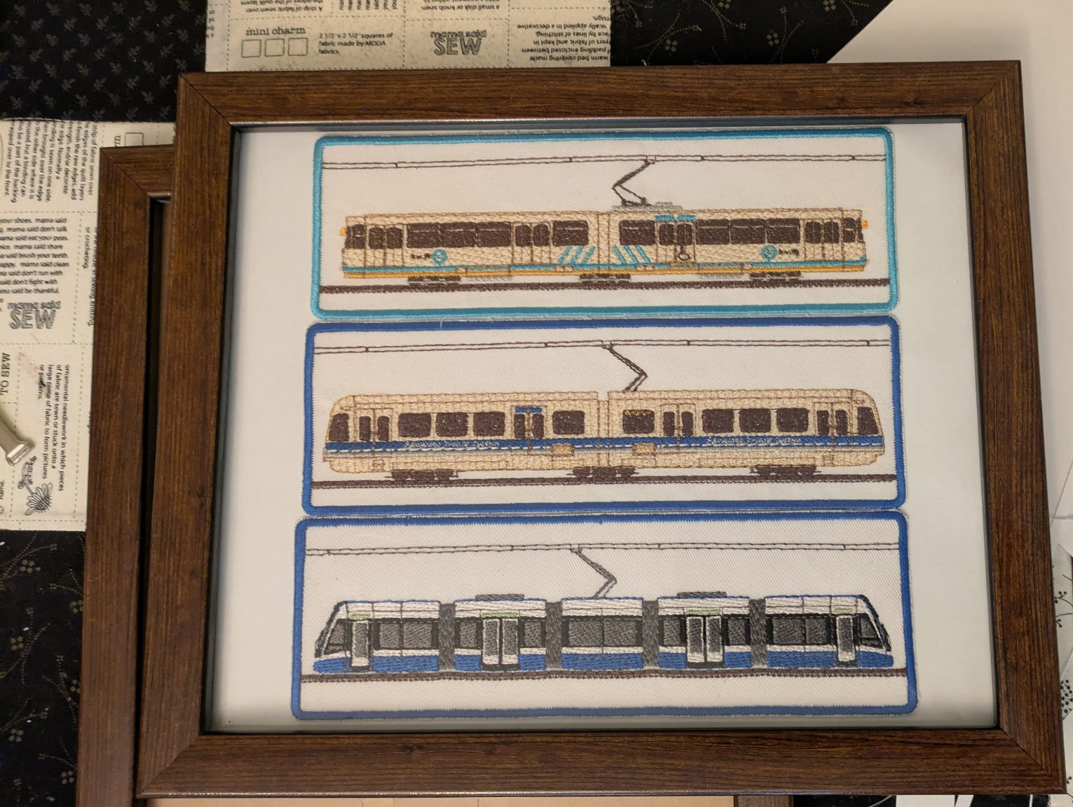 A photograph of three embroidered LRT train patches in a wooden frame