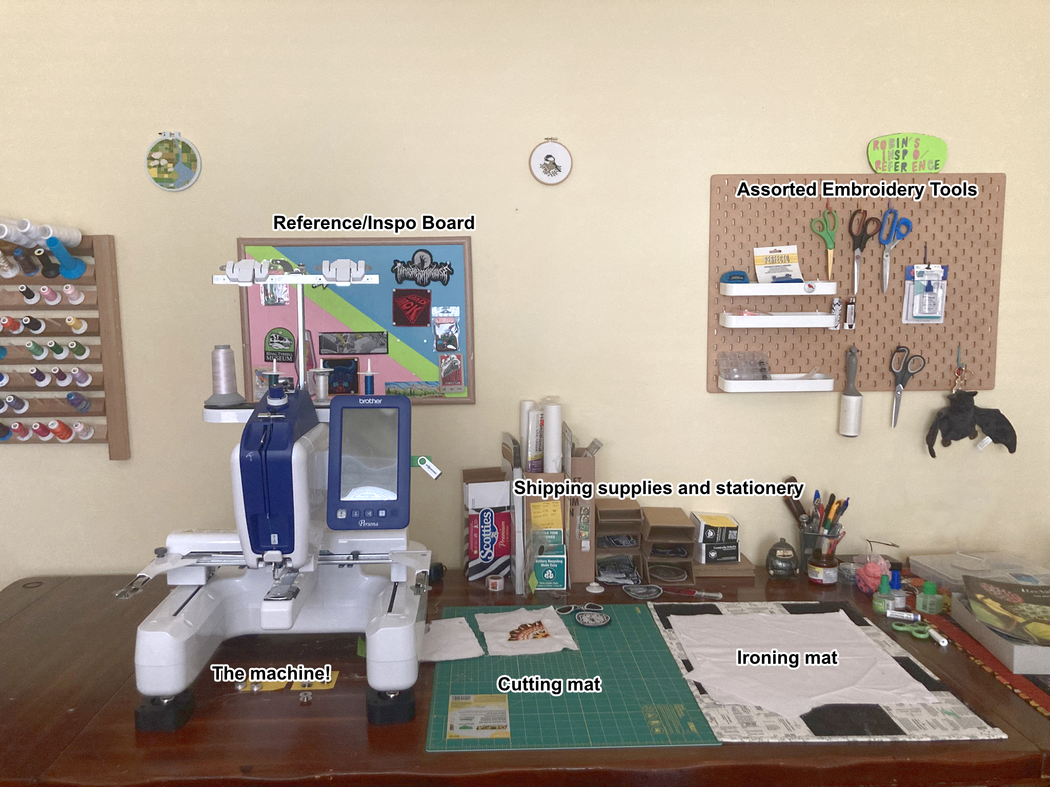 A photograph of the embroidery machine workspace. Several objects are shown, including the embroidery machine, a cutting and ironing mat, a pegboard with embroidery tools, and stationery supplies